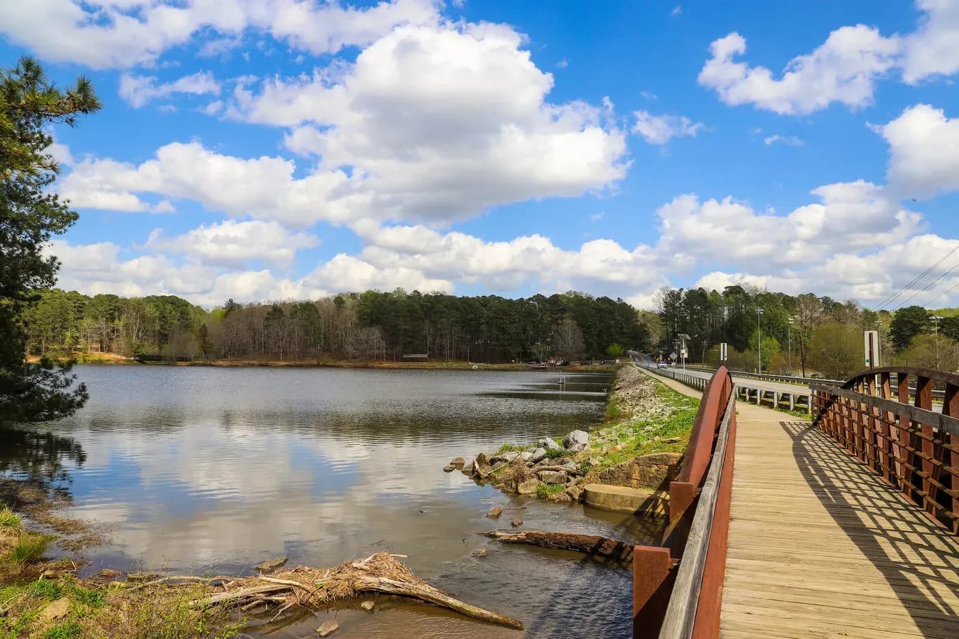 Wooden boardwalk along a calm lake with forested shoreline and blue sky with white clouds